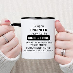 May include: A white ceramic mug with a black handle and rim, featuring black printed text that reads: Being an ENGINEER Is easy, it's like RIDING A BIKE EXCEPT THE BIKE IS ON FIRE YOU'RE ON FIRE EVERYTHING IS ON FIRE AND EVERYONE EXPECTS YOU TO FIX IT.