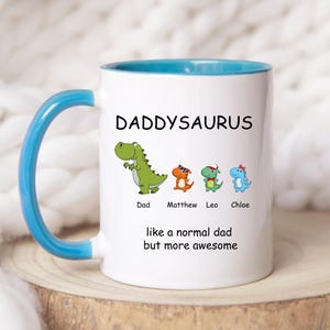 Puede incluir: Taza de café de cerámica blanca con borde y asa azules. La taza tiene un dinosaurio verde con el texto "Daddysaurus" encima. Debajo del dinosaurio hay cuatro dinosaurios más pequeños con los nombres "Dad", "Matthew", "Leo" y "Chloe" debajo. El texto "like a normal dad but more awesome" está impreso debajo de los dinosaurios.