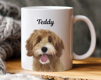 May include: White ceramic mug featuring a portrait of a dog with brown and tan fur, a pink tongue, and the name "Teddy" in black script. The mug has a curved handle and is set against a blurred background.