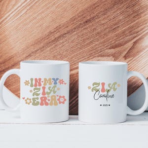 May include: Two white ceramic mugs featuring colourful graphic designs. The left mug displays the text IN MY ZIA ERA in wavy, multi-coloured letters with flower and smiley face icons. The right mug features the name Caroline and the year 2025.