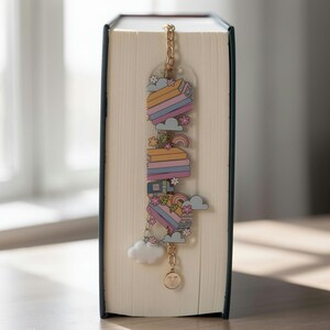 May include: A clear acrylic bookmark featuring a stack of colourful books, small flowers, rainbows, and clouds. The bookmark is attached to a gold-toned metal chain with a small white cloud charm and a round gold charm engraved with the letter V.