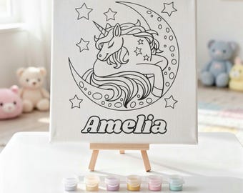 Puede incluir: Un lienzo cuadrado con una ilustración de líneas negras de un unicornio descansando sobre una luna creciente rodeada de estrellas. El nombre Amelia está impreso en letras negritas y delineadas debajo de la luna. Un caballete de madera sostiene el lienzo, con seis pequeños botes de pintura y un pincel azul colocados delante.