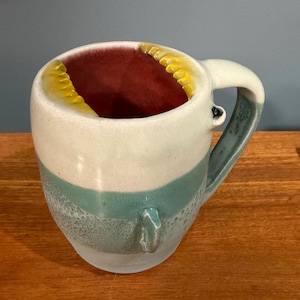 May include: A handmade ceramic mug with a unique design. The mug features a white top, a teal middle section, and a speckled bottom. The interior is a deep red, with yellow accents around the rim. The handle is teal and white.