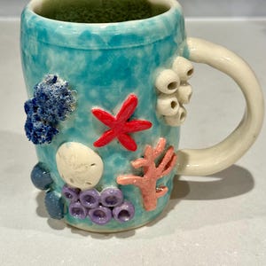 May include: A ceramic mug with a turquoise glaze, adorned with raised coral and sea life details. Features a red starfish, white seashells, and coral-like structures in shades of pink, blue, and purple. The handle is off-white, and the interior is green.