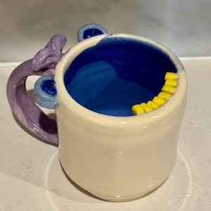 May include: A handmade ceramic mug with a cream-colored exterior and a vibrant blue interior. The handle is a lavender color with a unique, sculpted design. The rim features a row of yellow, tooth-like embellishments, adding a playful touch to the design.