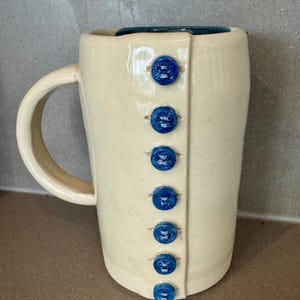 May include: A cream-coloured ceramic mug with a handle, featuring seven bright blue buttons down the front. The interior rim is teal. The mug has a simple, elegant design, suitable for tea or coffee.