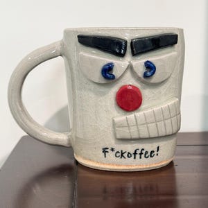 May include: A white ceramic coffee mug with a face sculpted on the front. The face has black eyebrows, blue eyes, a red nose, and a closed mouth. The mug has the text "F*ckoffee!" written on the bottom.