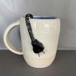 May include: A white ceramic mug with a black electrical plug design. The mug has a light beige base and a blue interior. The handle is on the left side. The plug is attached to the rim of the mug.