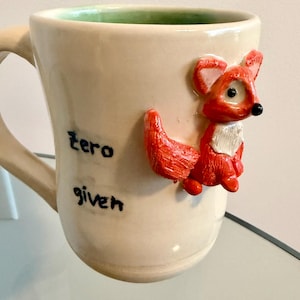 May include: Cream-colored ceramic mug with a 3D red fox figurine attached. The mug has the words "zero given" in black lettering. The interior of the mug is green.
