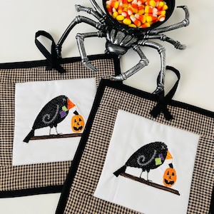 May include: Two Halloween-themed pot holders with a black crow holding a pumpkin, set against a white background and a brown and black checkered border. A black spider-shaped candy dish filled with candy corn sits above.