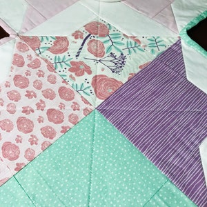 K&ouml;nnte beinhalten: Ein sternf&ouml;rmiger Patchwork-Decken mit verschiedenen floralen und geometrischen Mustern in Rosa-, Lila-, Gr&uuml;n- und Wei&szlig;t&ouml;nen.