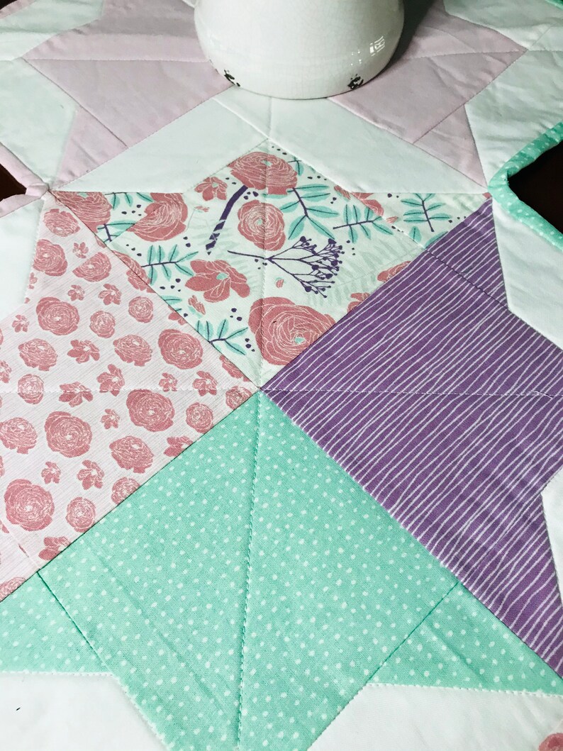 K&ouml;nnte beinhalten: Ein sternf&ouml;rmiger Patchwork-Decken mit verschiedenen floralen und geometrischen Mustern in Rosa-, Lila-, Gr&uuml;n- und Wei&szlig;t&ouml;nen.
