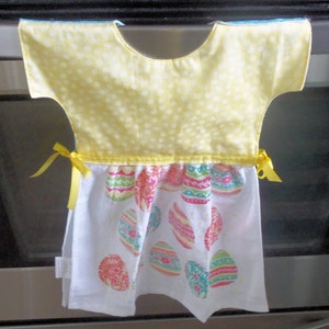 May include: A yellow and white kitchen towel with an Easter egg design. The top portion is yellow with white polka dots, and the bottom features colorful Easter eggs on a white background. Yellow ribbons are tied on each side.