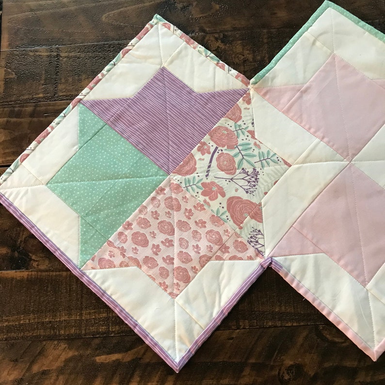 K&ouml;nnte beinhalten: Eine Patchwork-Decke mit wei&szlig;em Hintergrund und verschiedenen farbigen Quadraten. Die Quadrate sind in Rosa-, Gr&uuml;n- und Lilat&ouml;nen gehalten. Die Decke besteht aus verschiedenen Stoffen mit Blumen- und Streifenmustern.