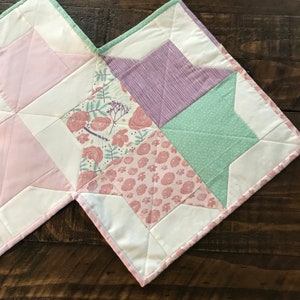 K&ouml;nnte beinhalten: Eine Patchwork-Decke mit einem Sternmuster in Rosa-, Gr&uuml;n- und Lilat&ouml;nen. Die Decke besteht aus verschiedenen Stoffen mit floralen und geometrischen Mustern.