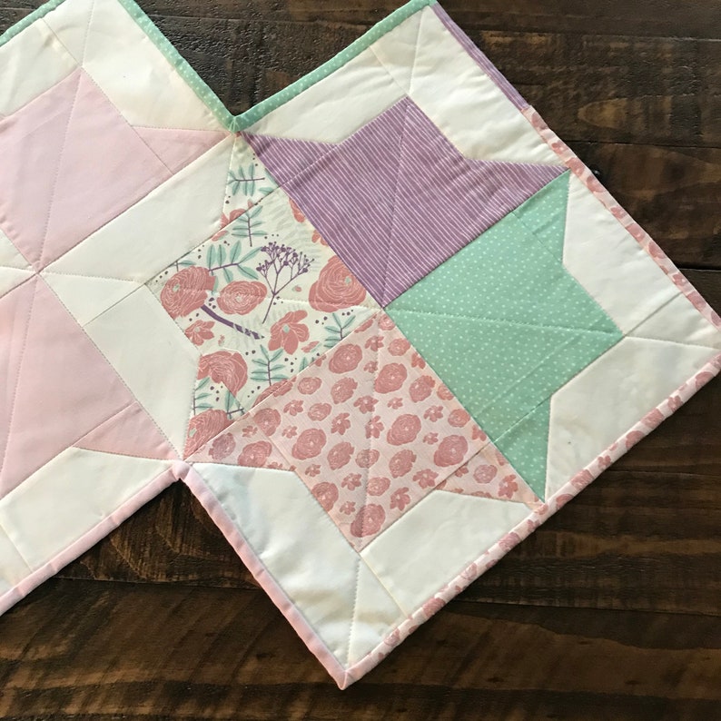 K&ouml;nnte beinhalten: Eine Patchwork-Decke mit einem Sternmuster in Rosa-, Gr&uuml;n- und Lilat&ouml;nen. Die Decke besteht aus verschiedenen Stoffen mit floralen und geometrischen Mustern.