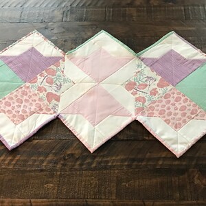 May include: A quilted table runner with a patchwork design featuring pink, green, and purple fabric squares. The runner has a white background and a floral pattern.