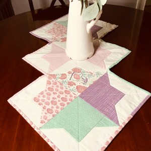 K&ouml;nnte beinhalten: Ein wei&szlig;er Tischl&auml;ufer mit Patchwork-Design aus rosa, gr&uuml;nem und violettem Stoff. Der L&auml;ufer ist mit einem Blumenmuster verziert und hat einen wei&szlig;en Rand. Ein wei&szlig;er Krug mit einem Blumenarrangement steht auf dem L&auml;ufer.