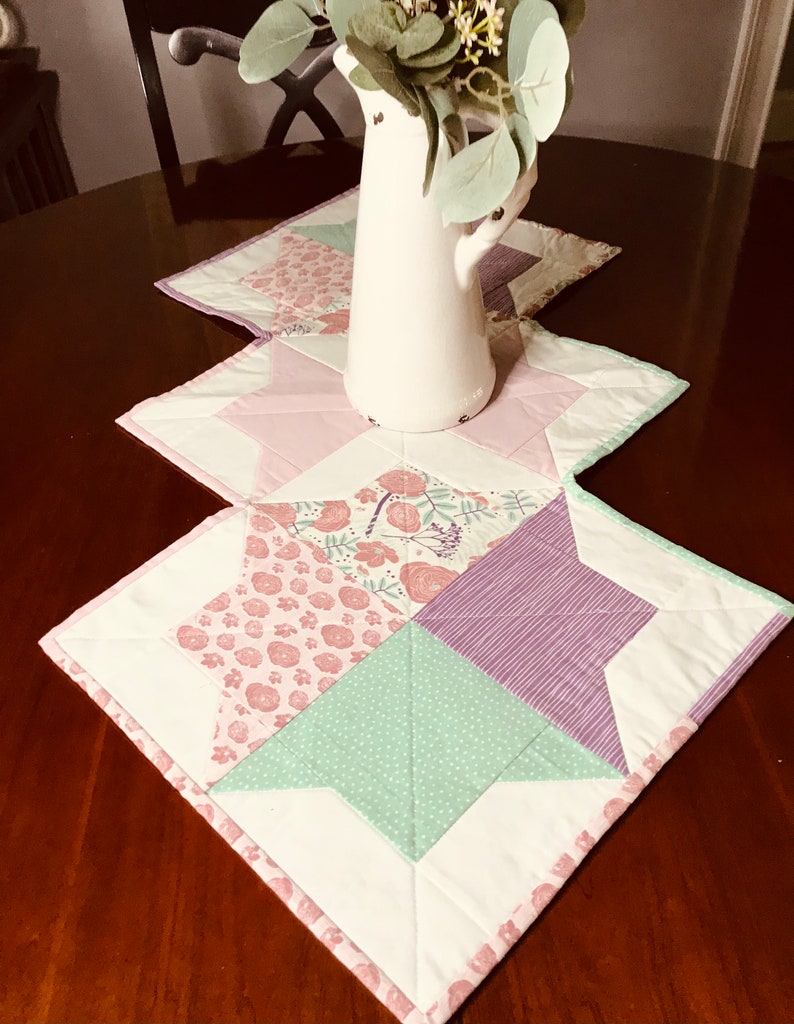 K&ouml;nnte beinhalten: Ein wei&szlig;er Tischl&auml;ufer mit Patchwork-Design aus rosa, gr&uuml;nem und violettem Stoff. Der L&auml;ufer ist mit einem Blumenmuster verziert und hat einen wei&szlig;en Rand. Ein wei&szlig;er Krug mit einem Blumenarrangement steht auf dem L&auml;ufer.