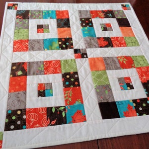 May include: A white table runner with a patchwork design featuring squares of various colors, including orange, brown, green, and blue. The squares are arranged in a geometric pattern, creating a visually appealing and modern look.