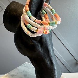 May include: Several colorful beaded bracelets in shades of pink, orange, white, and mint green are displayed on a black horse head sculpture. The bracelets have gold-colored spacer beads. The background is a light gray wall.