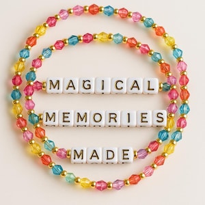 May include: Colorful beaded bracelets with the words "MAGICAL MEMORIES MADE" spelled out in white letter beads. The bracelets feature a variety of colors, including yellow, orange, pink, blue, and gold.