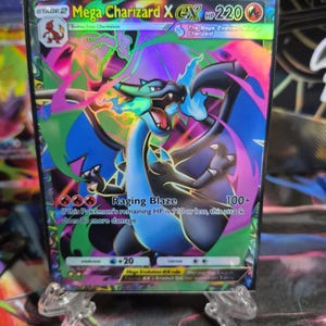 May include: A holographic Mega Charizard X EX Pokémon trading card. The card features a blue and black dragon with a fiery background of pink, green, and purple. The card text includes "Raging Blaze" and "HP 220".