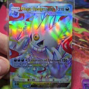 May include: A holographic Mega Gengar EX Pokémon card. The card features a white and purple Gengar with rainbow accents. Text includes "Mega Gengar EX", "Labyrinth of Shadows 120", and "HP 210".