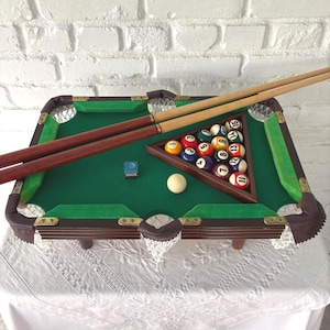 May include: A miniature pool table with a green felt surface, brown wooden frame, and corner pockets. Includes a set of numbered billiard balls, a white cue ball, two pool cues, and a wooden triangle rack. The table is set on a white lace tablecloth.