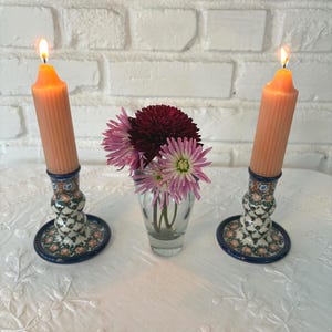 May include: Two fluted peach-colored taper candles burning in matching ceramic candle holders with a blue, green, and orange floral pattern, positioned on either side of a small clear glass vase containing dark red and light pink chrysanthemum flowers.