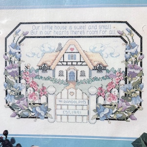 May include: Cross-stitch design of a quaint house with a thatched roof, surrounded by flowers and a white picket fence. The text reads "Our little house is sweet and small, but in our hearts there's room for all." The gate reads "The Randolphs, Est. 1991."