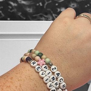 May include: Three beaded bracelets stacked on a wrist. The bracelets feature white letter beads spelling "REVIVAL" and "ZACH PINKSA", along with pastel-colored beads and a pink flower charm. The bracelets are a mix of colors including pink, green, and purple.