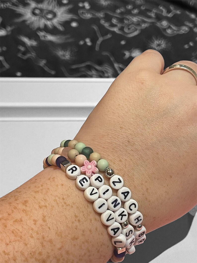May include: Three beaded bracelets stacked on a wrist. The bracelets feature white letter beads spelling "REVIVAL" and "ZACH PINKSA", along with pastel-colored beads and a pink flower charm. The bracelets are a mix of colors including pink, green, and purple.
