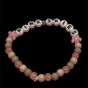 May include: A beaded bracelet with the word "PINK SKIES" spelled out in white letter beads. The bracelet features pink, beige, and green beads, with pink flower accents and a small charm. The background is black.