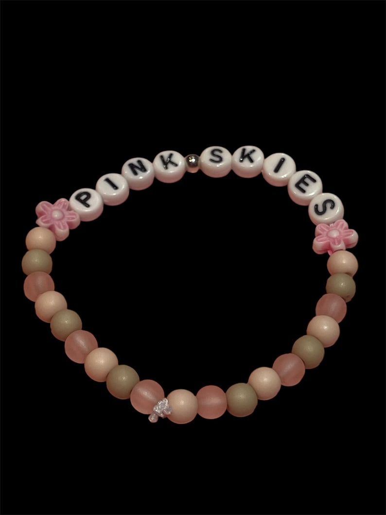 May include: A beaded bracelet with the word "PINK SKIES" spelled out in white letter beads. The bracelet features pink, beige, and green beads, with pink flower accents and a small charm. The background is black.
