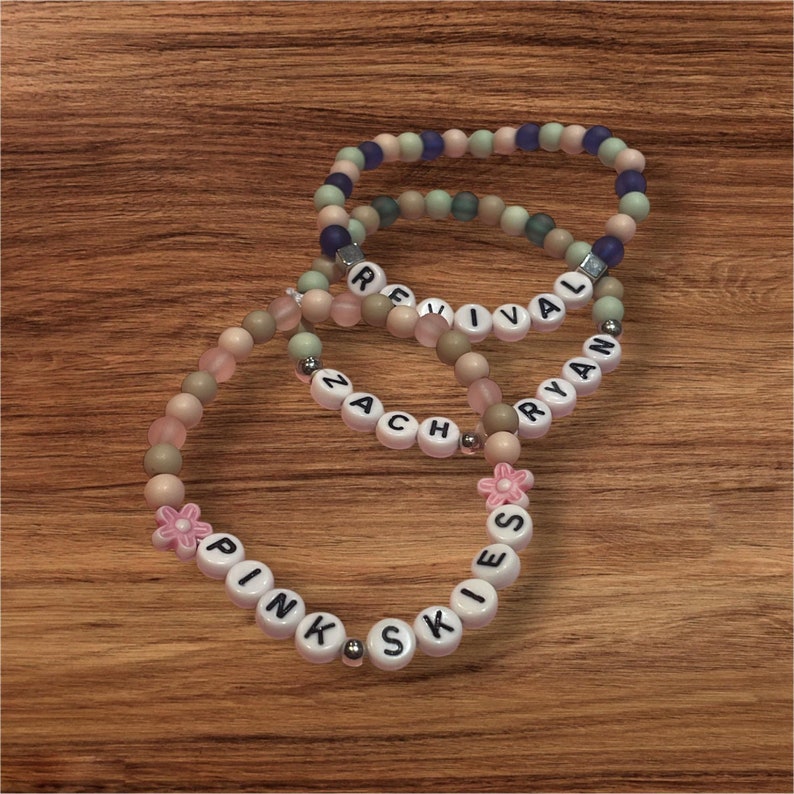 May include: Three beaded bracelets with pastel-colored beads and white letter beads spelling out "REVIVAL", "ZACH RYAN", and "PINK SKIES". One bracelet has a pink flower charm. The bracelets are on a wooden surface.