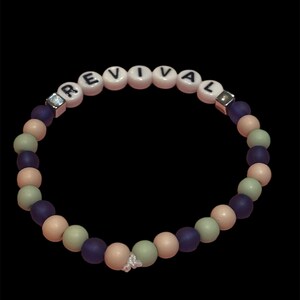 May include: A beaded bracelet with the word "REVIVAL" spelled out in white letter beads. The bracelet features a mix of purple, peach, and green beads, with a small butterfly charm. The bracelet is set against a black background.