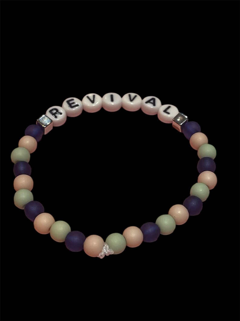 May include: A beaded bracelet with the word "REVIVAL" spelled out in white letter beads. The bracelet features a mix of purple, peach, and green beads, with a small butterfly charm. The bracelet is set against a black background.