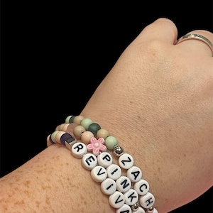 May include: Three beaded bracelets with white letter beads spelling "REVIVAL" and "ZACHS". The bracelets feature a variety of pastel-colored beads, including pink, green, and purple. A small pink flower bead accents one bracelet.