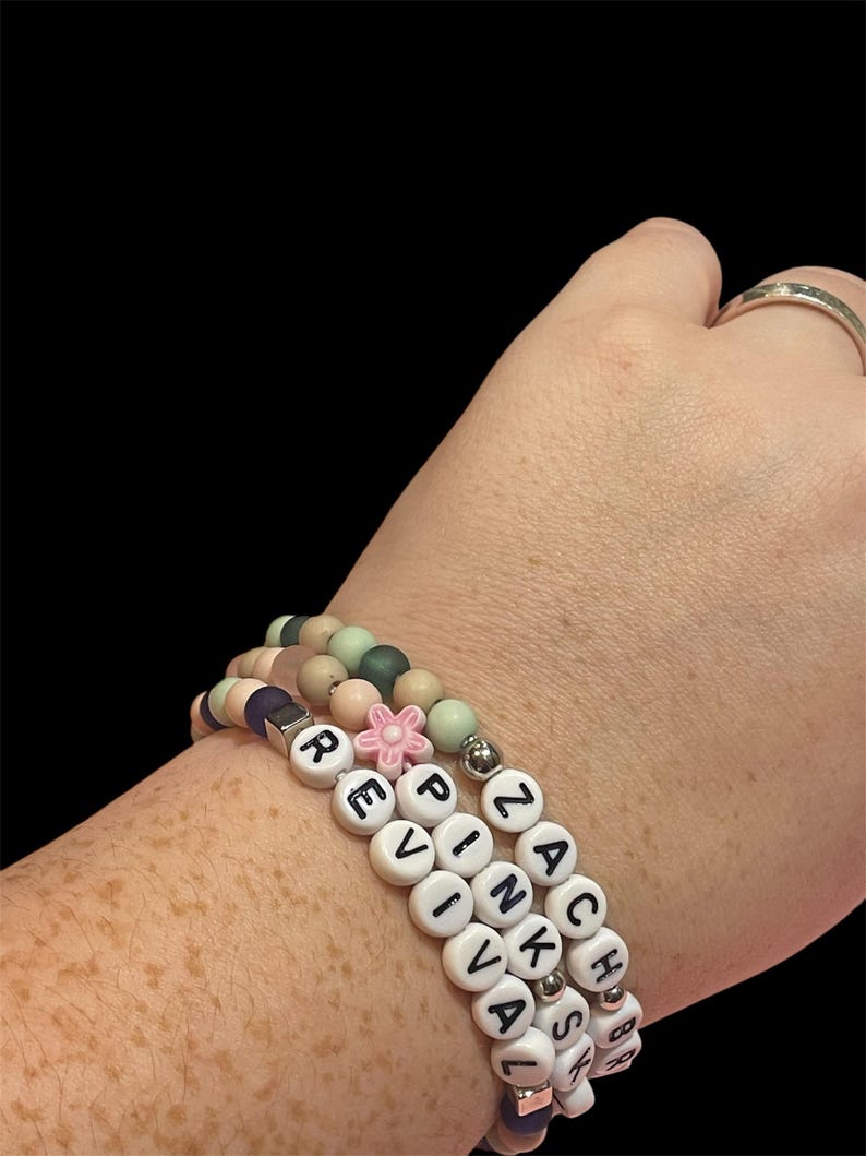 May include: Three beaded bracelets with white letter beads spelling "REVIVAL" and "ZACHS". The bracelets feature a variety of pastel-colored beads, including pink, green, and purple. A small pink flower bead accents one bracelet.