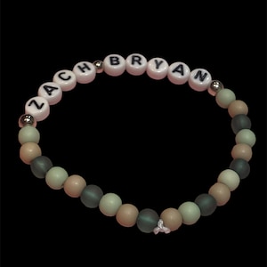 May include: A beaded bracelet with the name "ZACH BRYAN" spelled out in white letter beads on a pink background. The bracelet features a mix of matte gray, tan, and light green beads, with silver accents and a small charm. The bracelet is set against a black background.