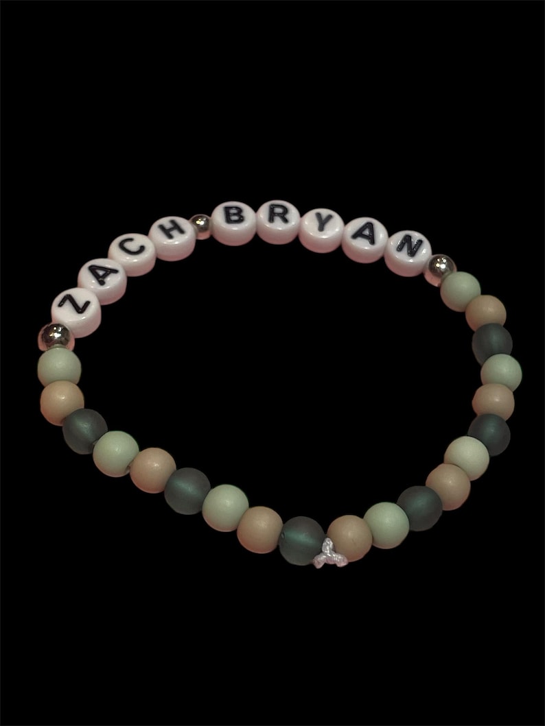 May include: A beaded bracelet with the name "ZACH BRYAN" spelled out in white letter beads on a pink background. The bracelet features a mix of matte gray, tan, and light green beads, with silver accents and a small charm. The bracelet is set against a black background.