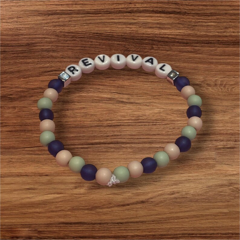 May include: A beaded bracelet with the word "REVIVAL" spelled out in white letter beads. The bracelet features a mix of blue, pink, and green beads, with a small white charm. The bracelet is displayed on a wooden surface.