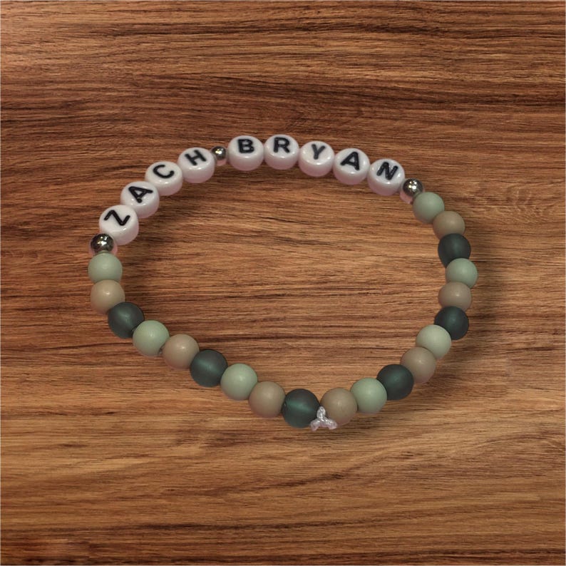 May include: A beaded bracelet with the name "ZACH BRYAN" spelled out in white letter beads. The bracelet features a mix of green, tan, and dark green beads. The bracelet is on a wooden surface.