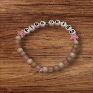 May include: A beaded bracelet with the word "PINK SKIES" spelled out in white letter beads. The bracelet features pink, beige, and green beads, along with pink flower-shaped accents. A small silver-colored bead is also present.