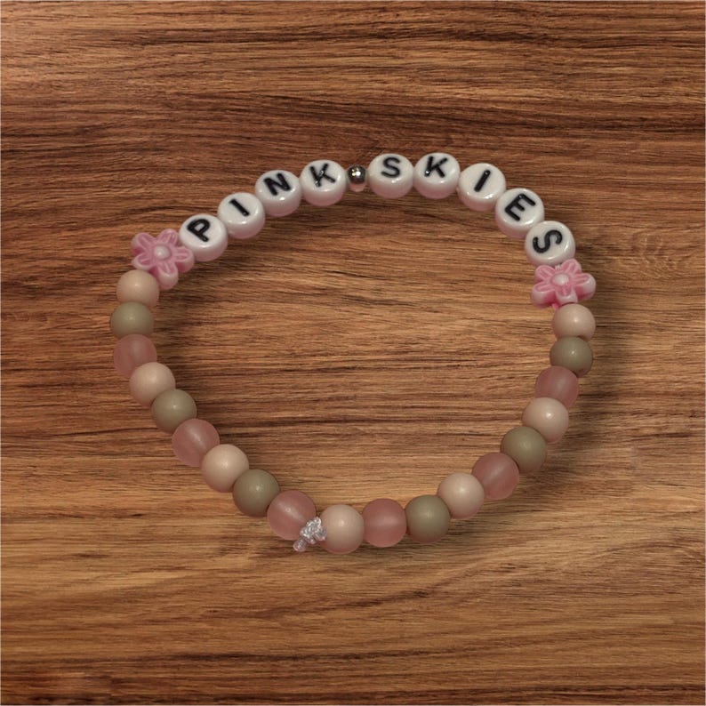 May include: A beaded bracelet with the word "PINK SKIES" spelled out in white letter beads. The bracelet features pink, beige, and green beads, along with pink flower-shaped accents. A small silver-colored bead is also present.