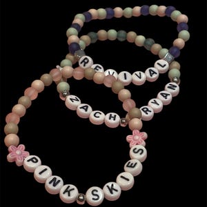 May include: Three beaded bracelets with white letter beads spelling "REVIVAL", "ZACH", and "PINK SKIES". The bracelets feature a mix of pink, beige, green, and purple beads, with a pink flower accent on one bracelet. The background is black.