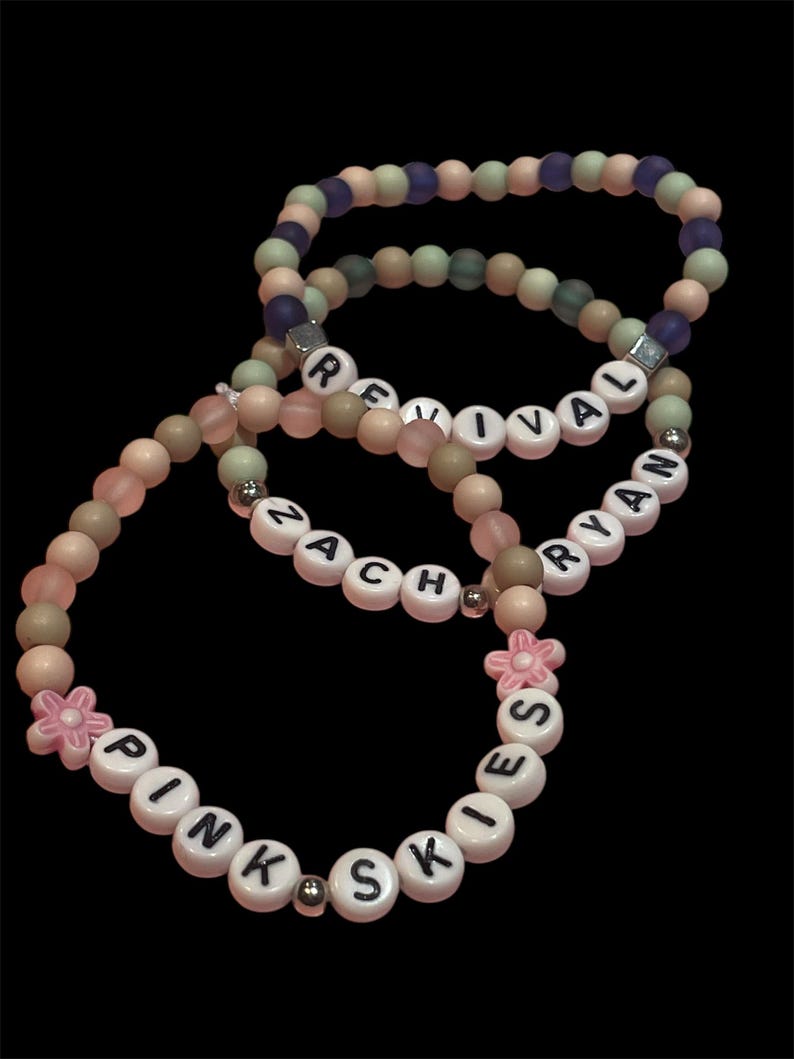 May include: Three beaded bracelets with white letter beads spelling "REVIVAL", "ZACH", and "PINK SKIES". The bracelets feature a mix of pink, beige, green, and purple beads, with a pink flower accent on one bracelet. The background is black.