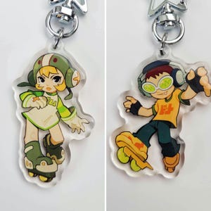 May include: Two colorful acrylic keychains featuring cartoon characters on roller skates. The keychain on the left features a female character with blonde hair wearing a green and white outfit and a helmet. The keychain on the right features a male character with brown hair wearing a yellow and green outfit and headphones.