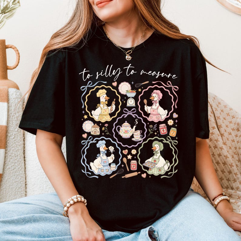 May include: Black t-shirt featuring a whimsical design with the text "to silly to measure." The graphic includes cartoon geese in chef hats and aprons, surrounded by baking-related illustrations. The design is in pastel colors.